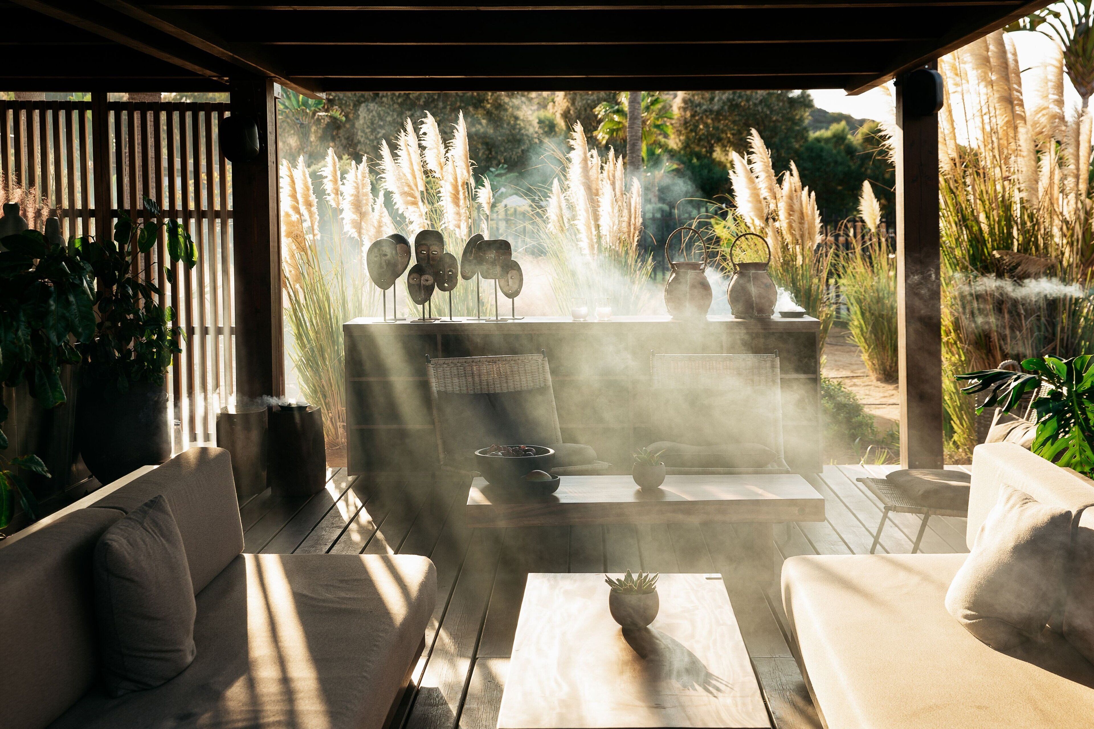 A covered outdoor seating area with a fire feature, surrounded by plants and a view of tall grasses.