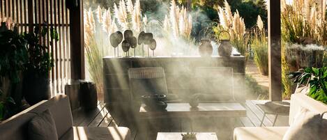 A covered outdoor seating area with a fire feature, surrounded by plants and a view of tall grasses.