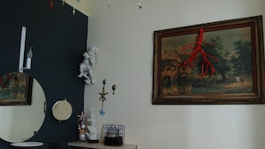 A room with a dark blue wall, a round mirror, a small shelf with decorative items, and a framed painting with a red tree branch. - Kemmare (Riposto)