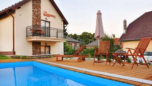 Una piscina con sillas de madera y una mesa, un edificio con balcón y una chimenea de ladrillo.