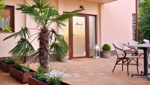 Entrada de un hotel con mostrador de recepción, una palmera, plantas en macetas y asientos al aire libre.
