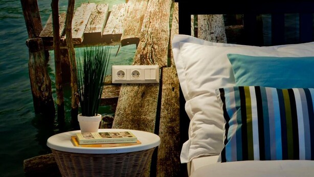 Un dormitorio con un muelle de madera que se adentra en un cuerpo de agua, una lámpara y una pequeña mesa con una planta y un libro.