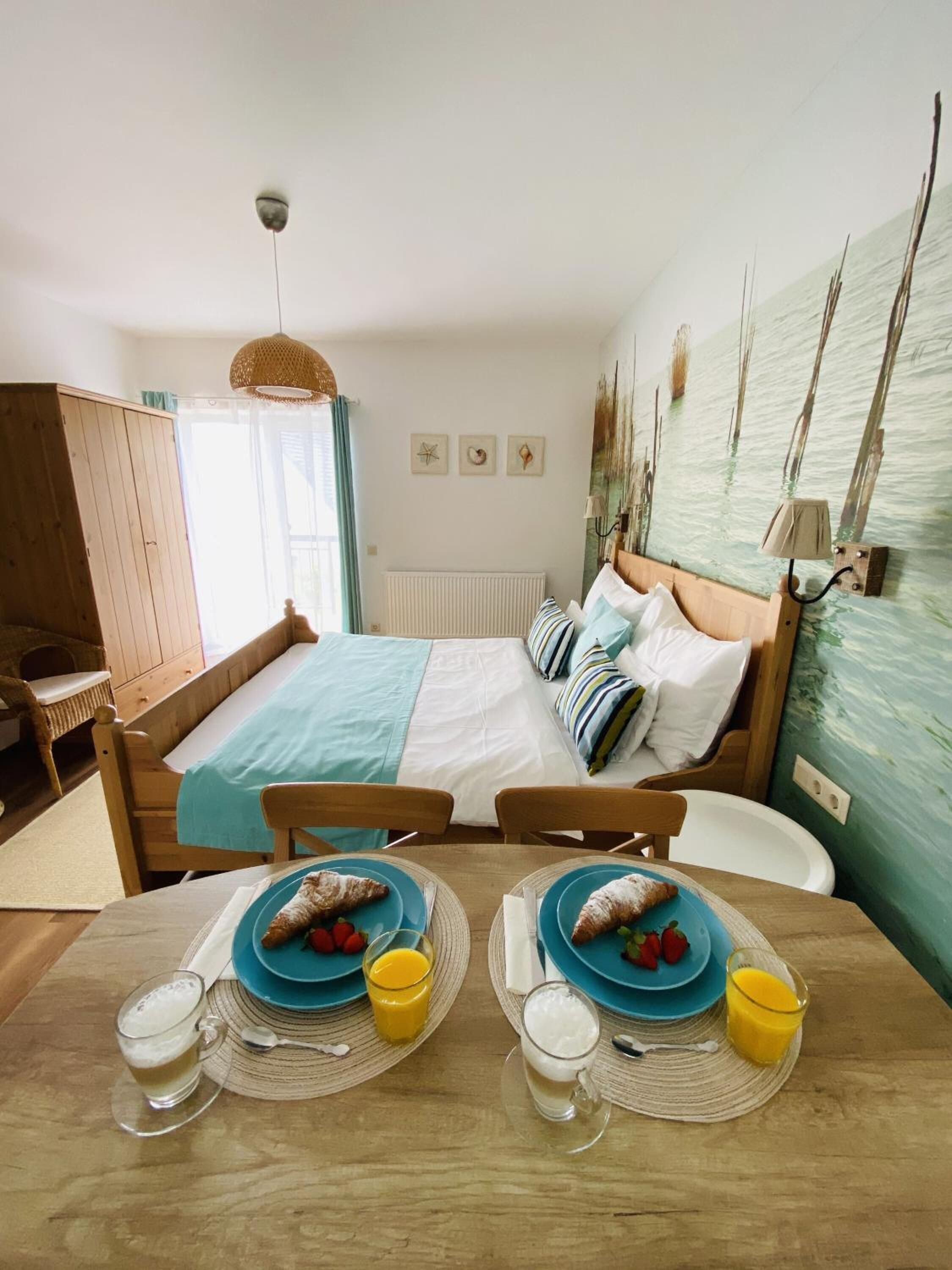 A bedroom with a bed, a wooden wardrobe, a dining table with breakfast plates, and a wall with a painting.