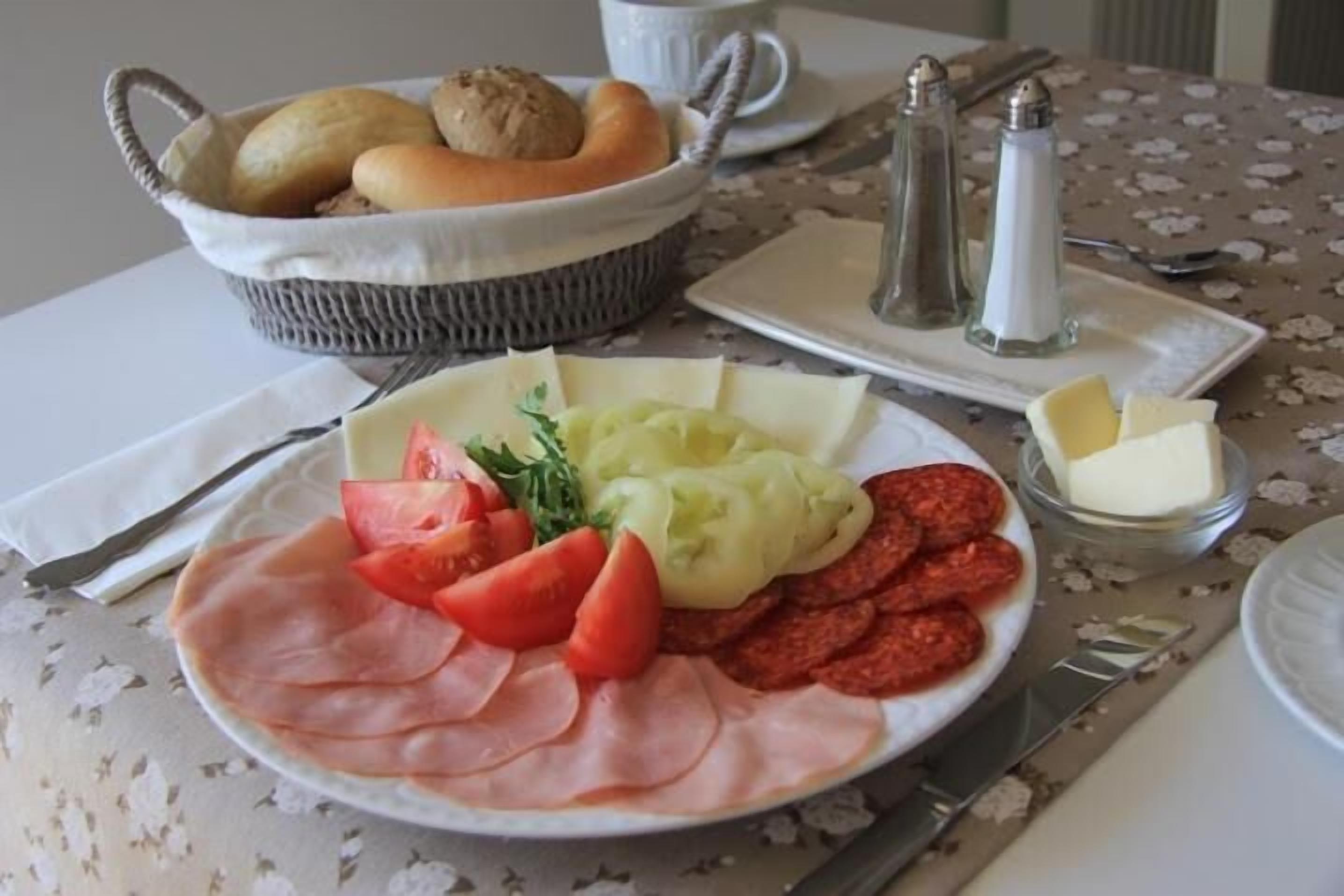 Un piatto con prosciutto a fette, pomodori e salsicce, accompagnato da una ciotola di pane e un piatto di purè di patate.