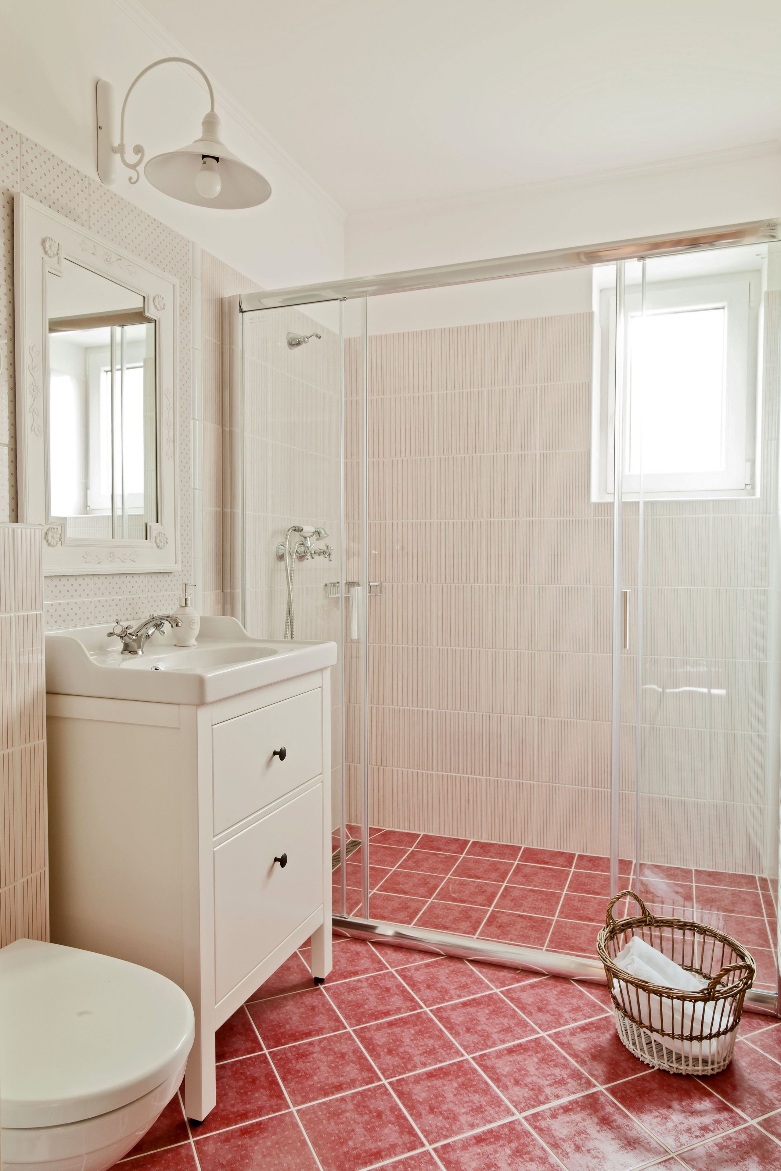A bathroom with a white sink, mirror, and toilet, a shower with a glass door, and a red-tiled floor.