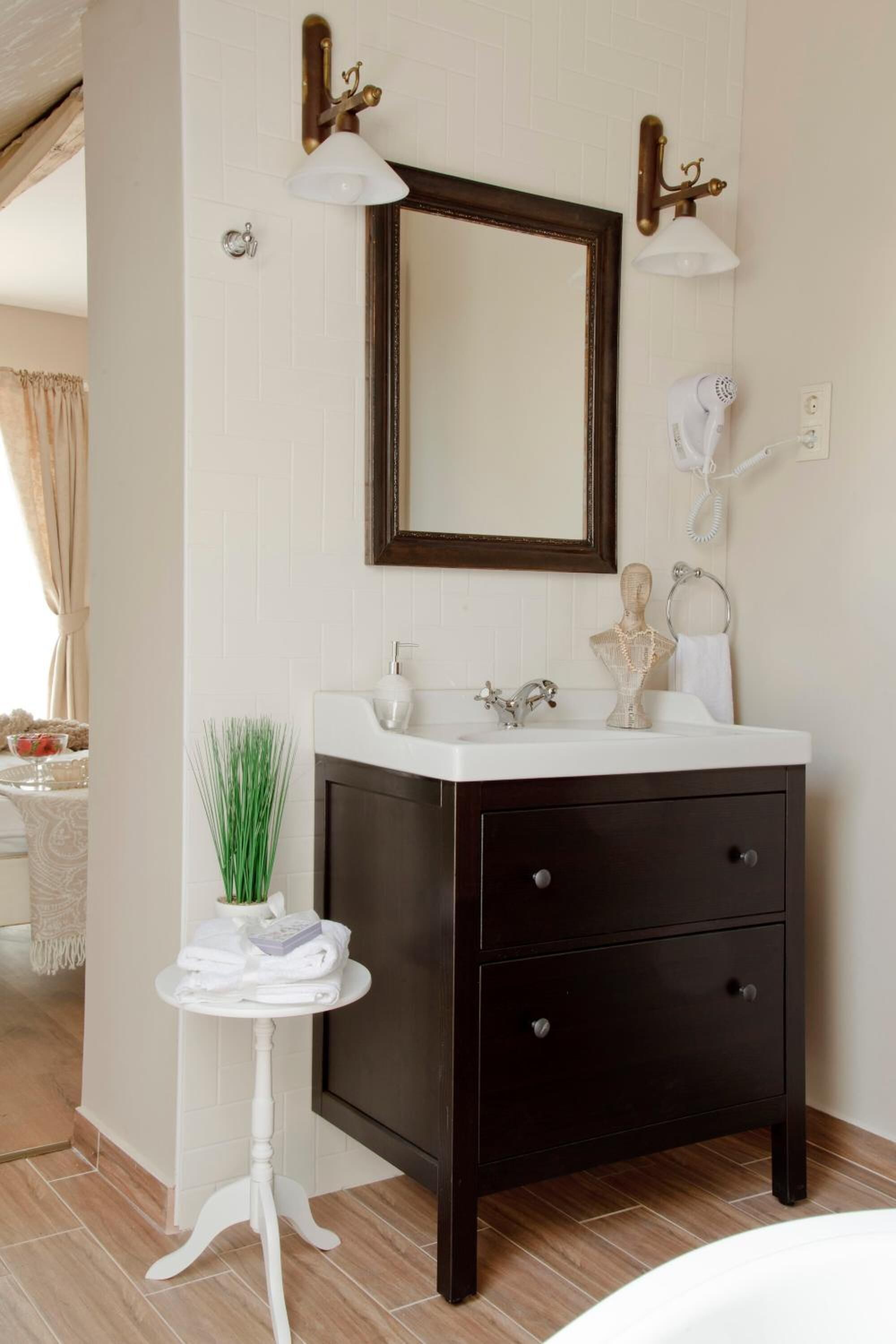A bathroom with a dark wooden vanity, white sink, and a mirror with a brass frame. There is a small white table with a plant and towels beside the vanity.