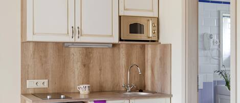 Una cocina compacta con armarios de madera, un microondas y un refrigerador pequeño. Una mesa de comedor con tazas y platos está dispuesta frente a la cocina.