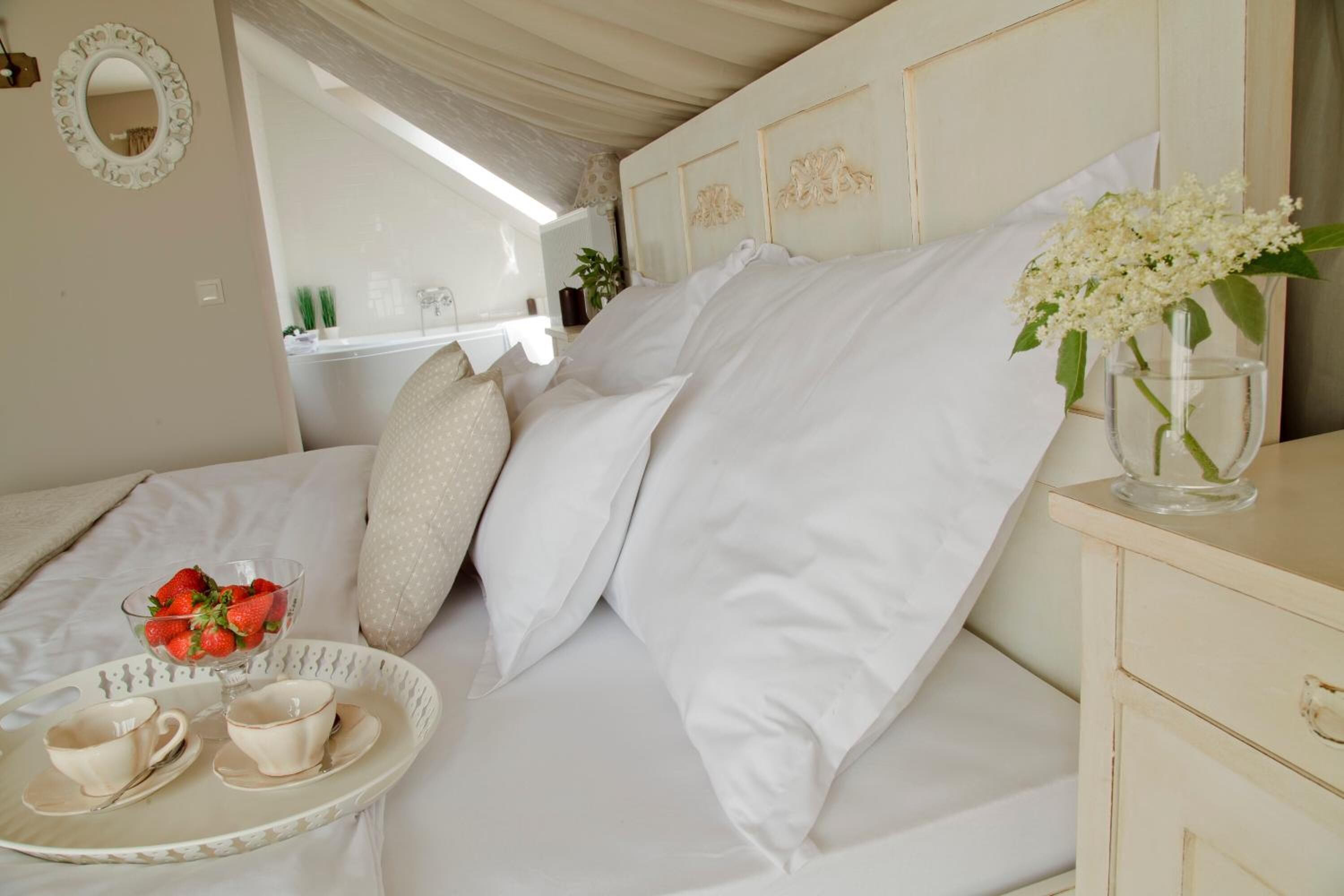 Una cama bien tendida con sábanas blancas, una bandeja con tazas y fresas, y un florero con flores sobre una mesita de noche.
