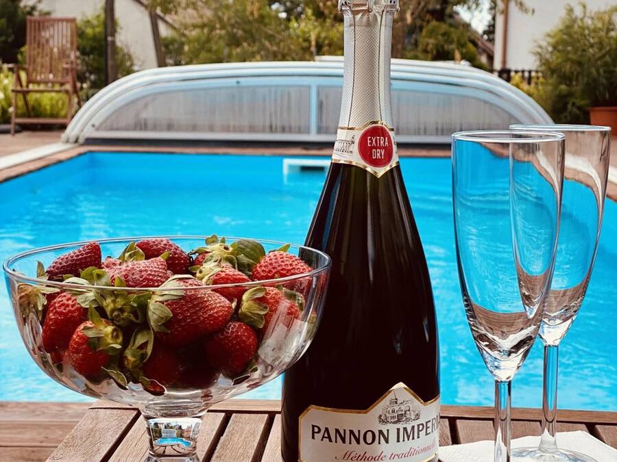 Une bouteille de vin Pannon Imperiale, un bol de fraises et deux verres à vin sur une table en bois, près d’une piscine.