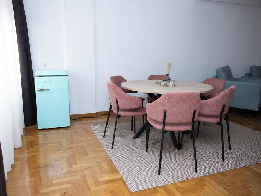 Un comedor pequeño con una mesa redonda, seis sillas rosas, un refrigerador verde azulado y un tapete estampado.