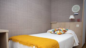 Una cama individual con una colcha blanca y un plaid amarillo mostaza, un cabecero de madera, una lámpara redonda fijada a la pared y una mesita de noche pequeña.