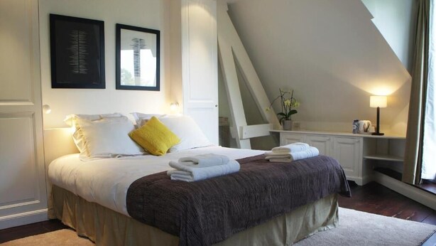 Un dormitorio con cama, almohadas, mesita de noche, lámpara y ventana.