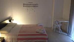 Una cama bien hecha con ropa de cama a rayas, una mesita pequeña y una silla en una habitación sencilla con una partitura musical colgada en la pared.