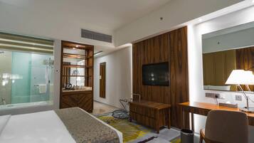 A modern hotel room with a bed, a desk, a TV, a mirror, and a shower area.