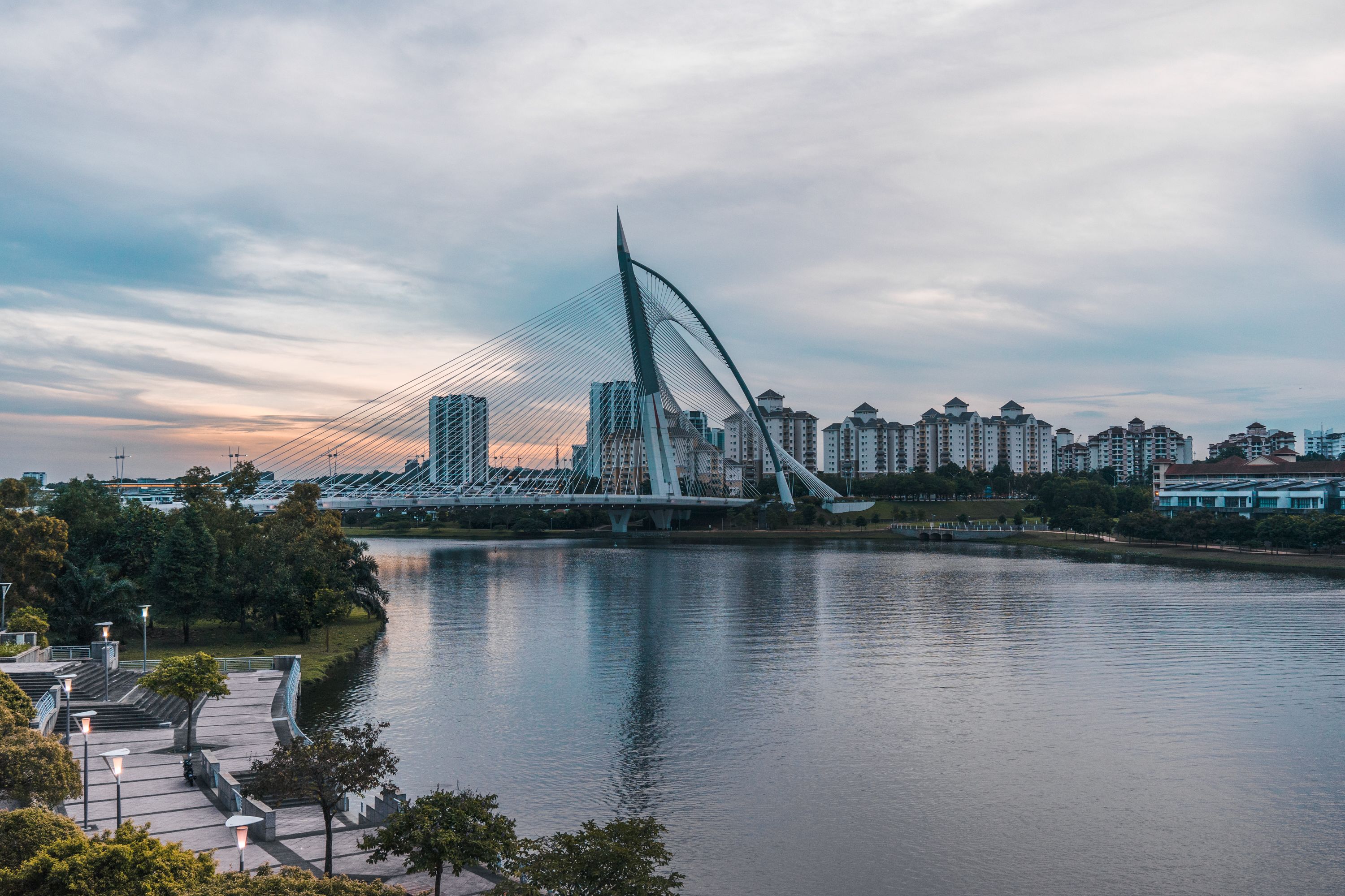 A modern bridge with a unique design spanning a river, surrounded by high-rise buildings and a park area with pathways and trees.
