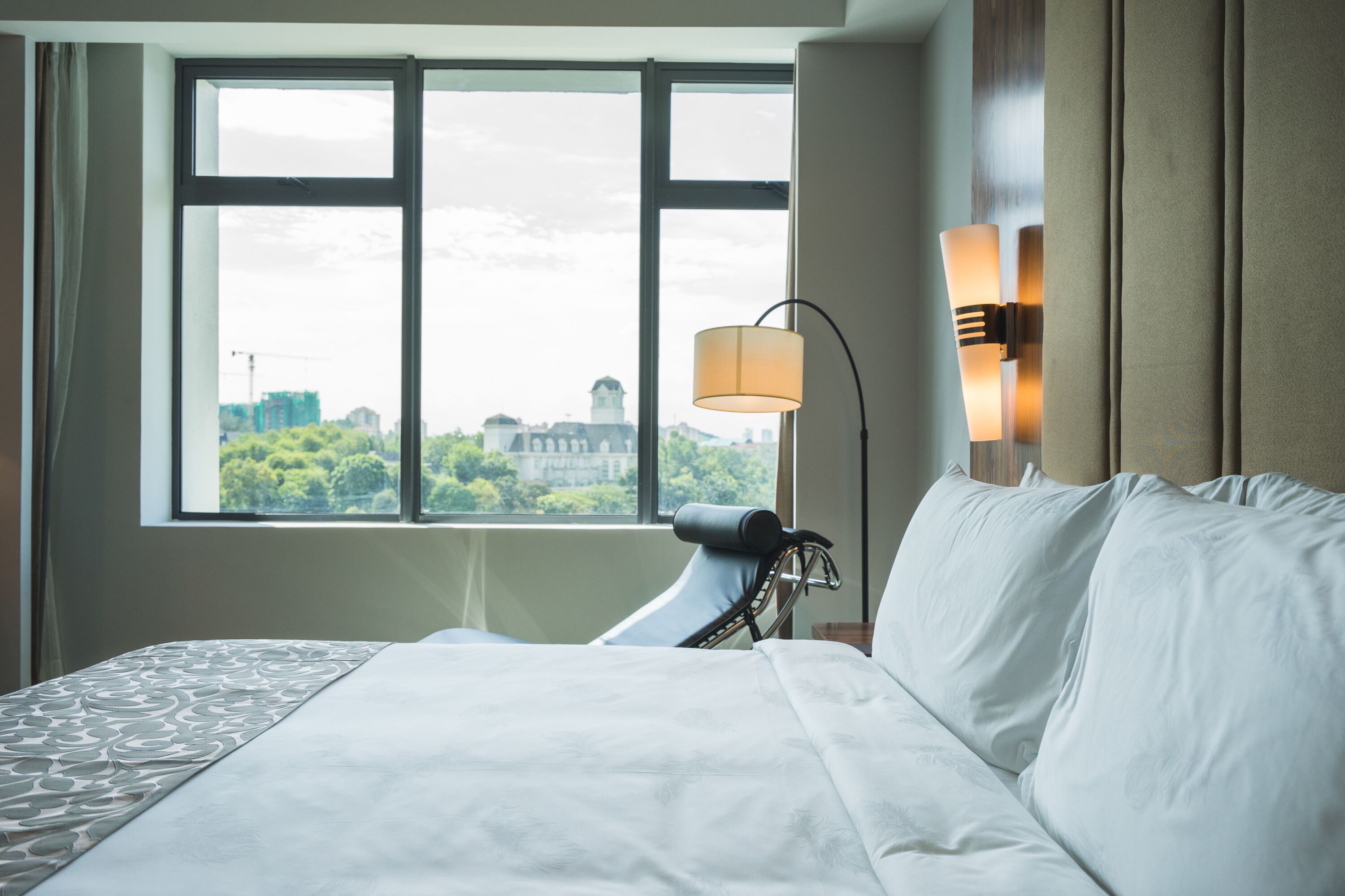Una habitación de hotel con una cama grande, una ventana que ofrece vistas a edificios y vegetación, y una lámpara fijada a la pared.