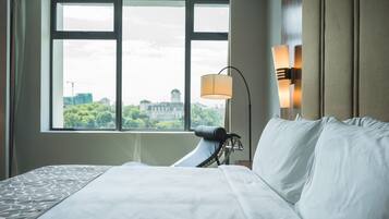 Una habitación de hotel con una cama grande, una ventana que ofrece vistas a edificios y vegetación, y una lámpara fijada a la pared.
