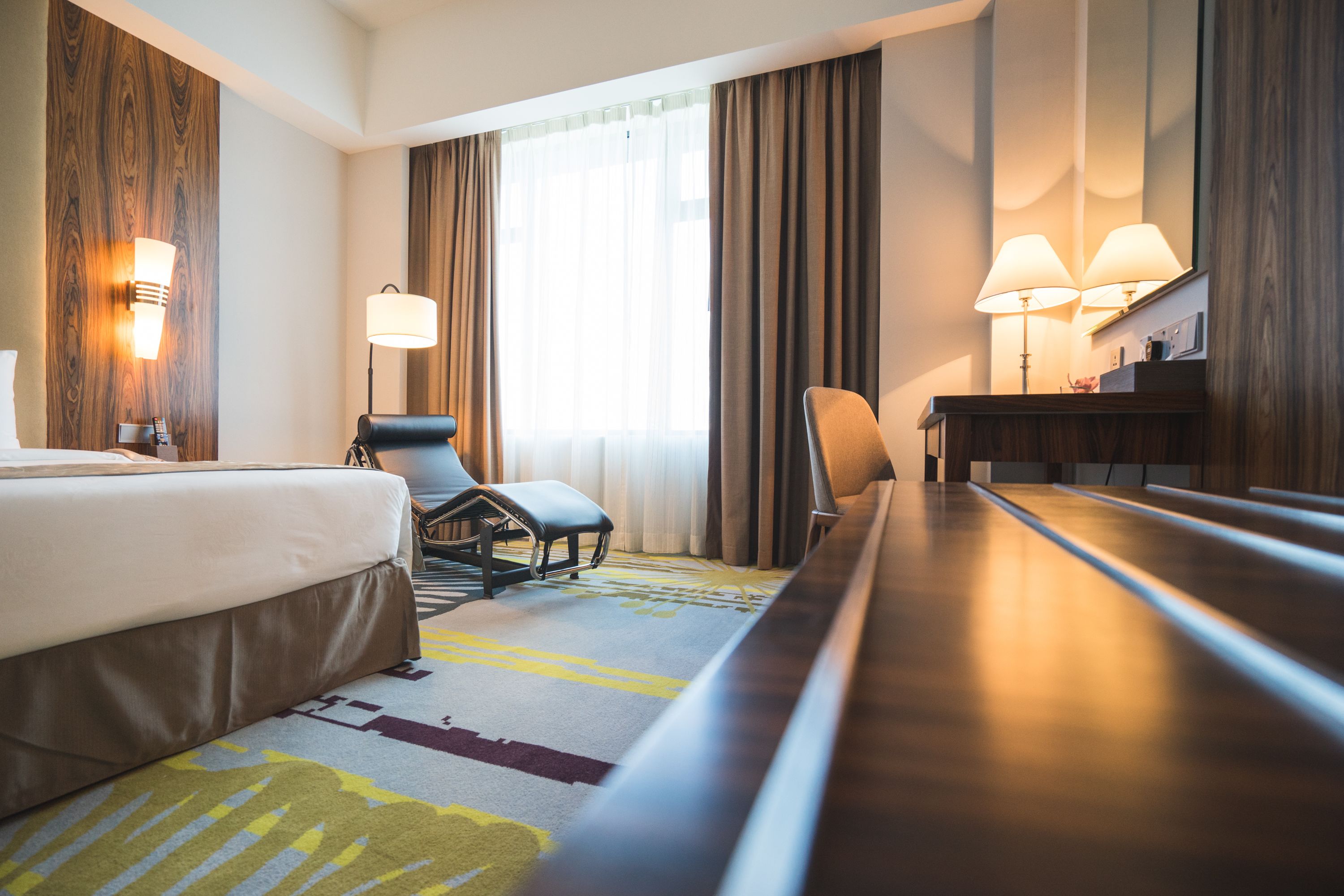 Une chambre d’hôtel avec un lit, deux chaises, un bureau et une grande fenêtre avec des rideaux.