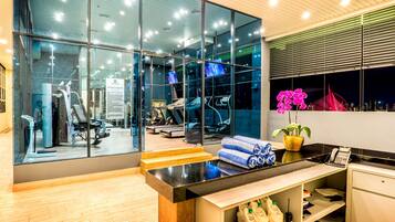A modern hotel lobby with large glass windows, a reception desk, and a view of the gym area.