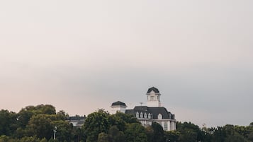 Blick auf das Wasser mit einem Steg, der ins ruhige Wasser führt, ein großes Gebäude mit einem Turm im Hintergrund und ein bewaldetes Ufergebiet.