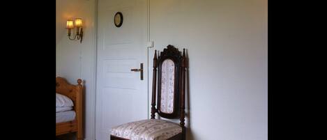 A room with a wooden bed, a chair with a floral cushion, a white door with a clock, and a wooden beam above the door.