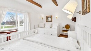 A white bedroom with a large bed, a window with curtains, a wooden ceiling beam, and a wall-mounted lamp. - Kullabygdens Bed & Breakfast (Jonstorp)