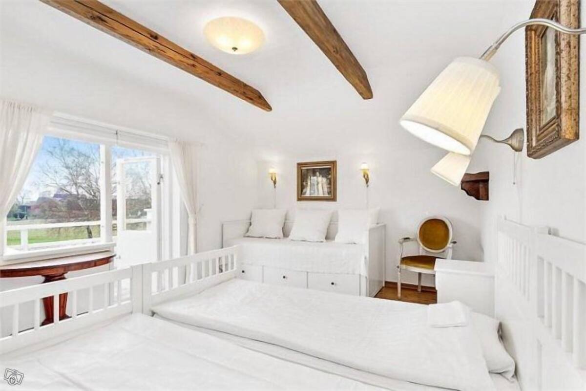 Un dormitorio blanco con una cama grande, una ventana con cortinas, una viga de techo de madera y una lámpara fijada a la pared.