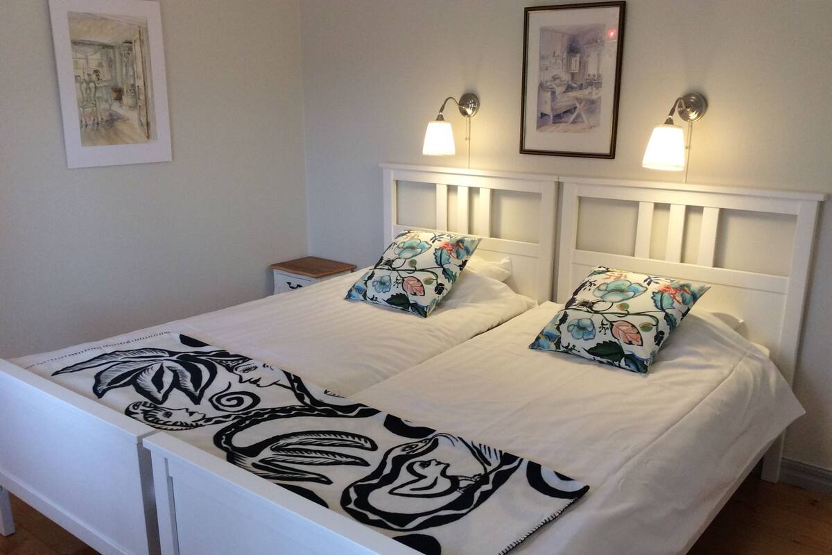 Una cama bien hecha con almohadas florales y una manta blanco y negro, una mesita de noche y lámparas fijadas a la pared.