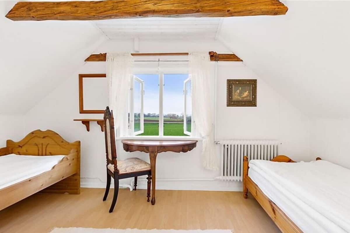 Un dormitorio con dos camas de madera, una silla de madera, una mesita redonda y un ventanal con vistas a un campo verde.