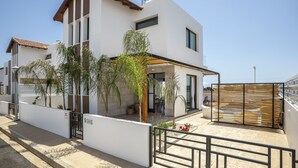 Una casa moderna de dos pisos con balcón, cerca viva de madera y plantas en macetas.