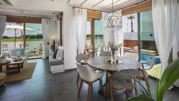 Una sala de estar moderna con una mesa de comedor redonda, sofás blancos y un amplio ventanal corredero que da a una zona de piscina.
