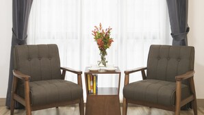 Una habitación con dos sillones, una mesita con un jarrón de flores y una ventana con cortinas ligeras.