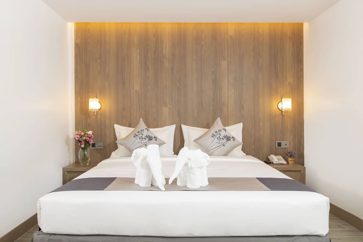 A modern hotel room with a wooden headboard, two bedside tables, a bed with white linens, decorative pillows, a vase with flowers, and a telephone on the nightstand.