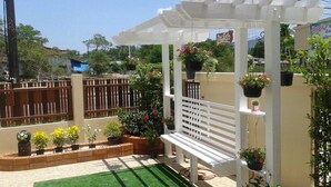 A covered patio area with a white bench, potted plants, and a pergola structure. - Baanmeerak at Ranong (Ranong)