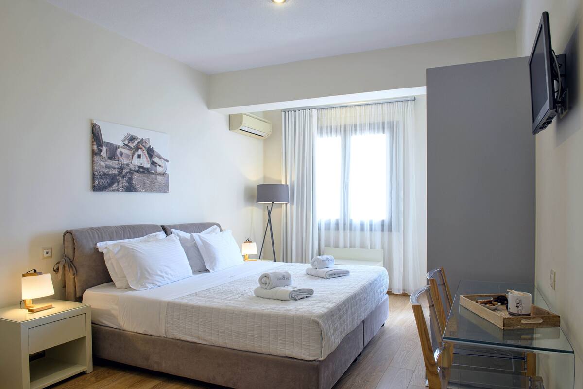 A hotel room with a bed, bedside table, lamp, TV, and a window with curtains.