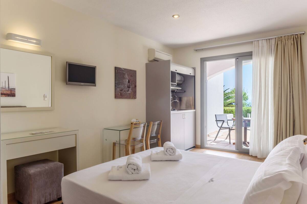 A hotel room with a bed, a desk, a chair, a TV, and a balcony with a table and chairs.