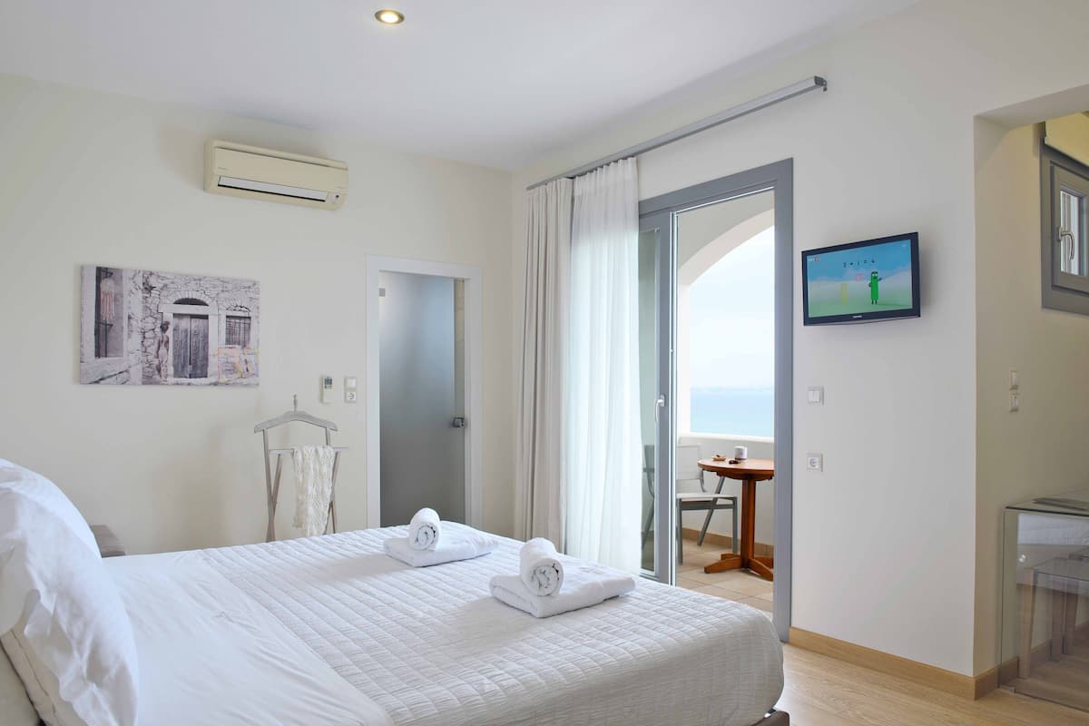 A hotel room with a bed, a TV, a painting, and a view of the sea.