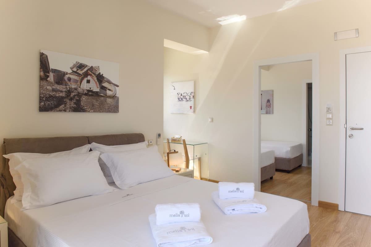 A hotel room with a bed, two pillows, a bedside table with a lamp, a painting on the wall, and a door leading to another room.
