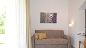 A living room with a beige sofa, a painting on the wall, and a table with a chair. - Melia Sol Art Studios (Chios)
