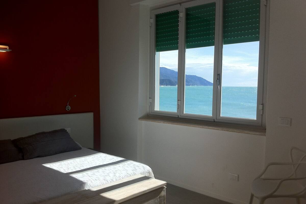 A bedroom with a bed, a window with blinds, a chair, and a view of the sea and mountains.