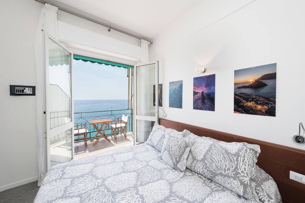 A bedroom with a bed, a balcony with a table and chairs, and a view of the sea.