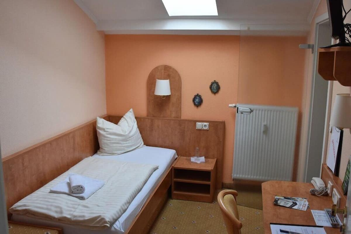 A hotel room with a bed, a desk, a chair, a lamp, a radiator, and a window.