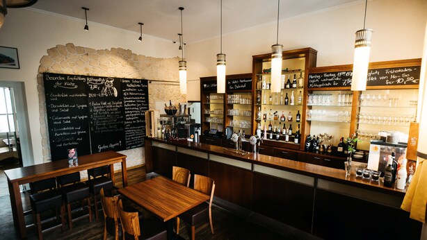 Un interior de restaurante con una mesa y sillas de madera, una zona de bar con botellas y vasos, y un menú en una pizarra.