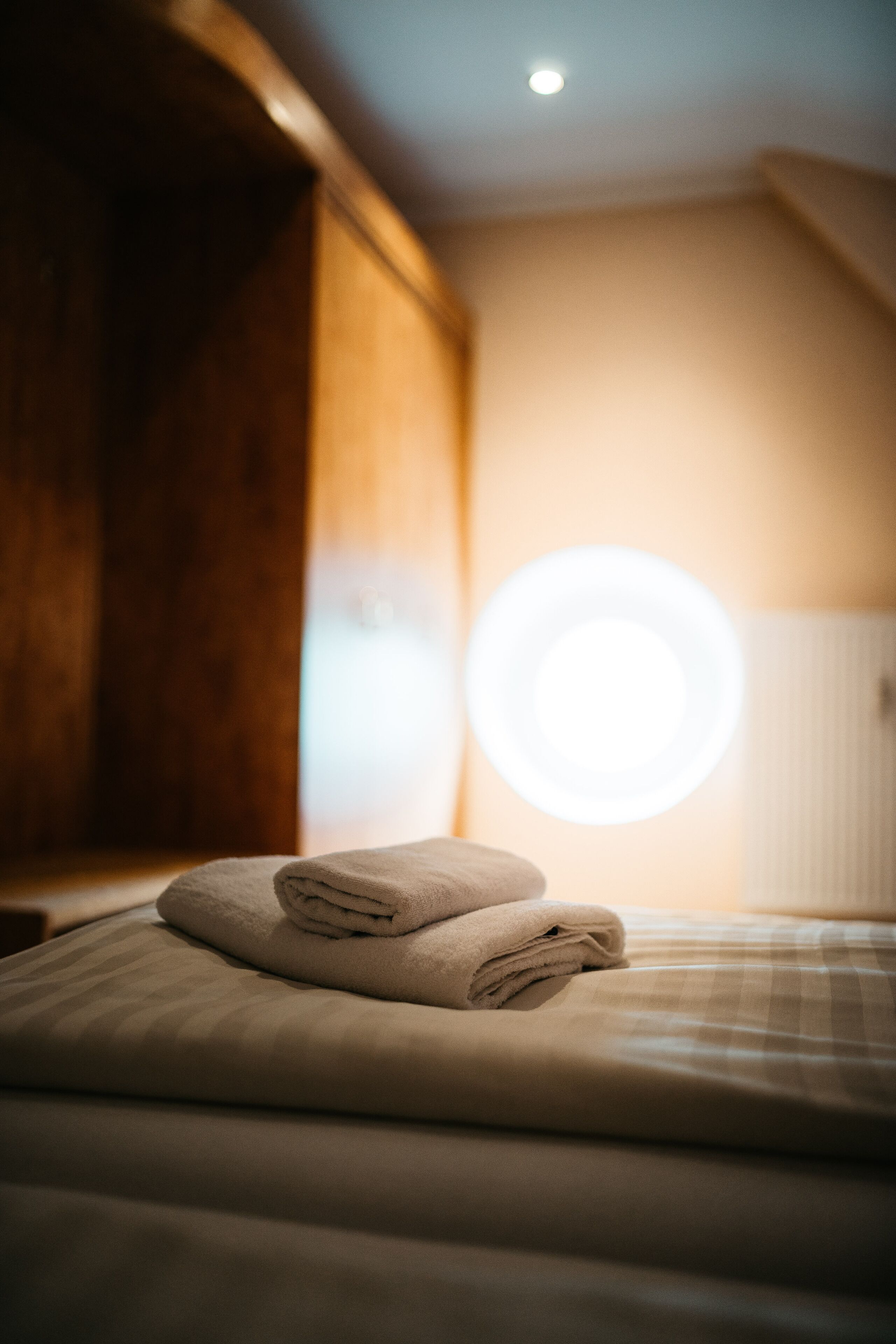 Una cama bien hecha con toallas dobladas y una lámpara circular al fondo.