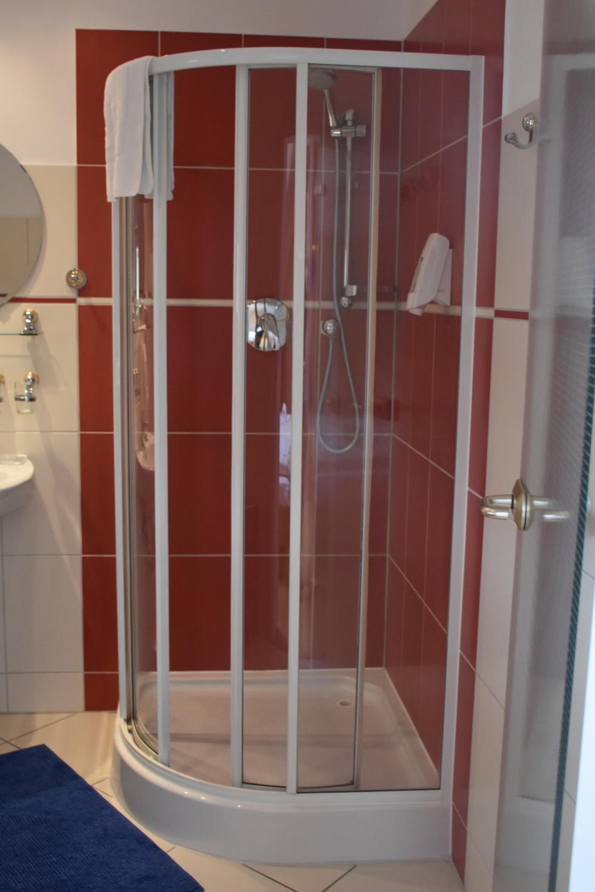 A glass-enclosed shower stall with a white door, red-tiled walls, a white sink, and a blue bath mat.