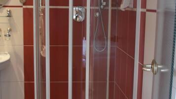 Un box de ducha con paredes de vidrio y puerta blanca, azulejos rojos y un lavamanos blanco, junto con un tapete de baño azul.