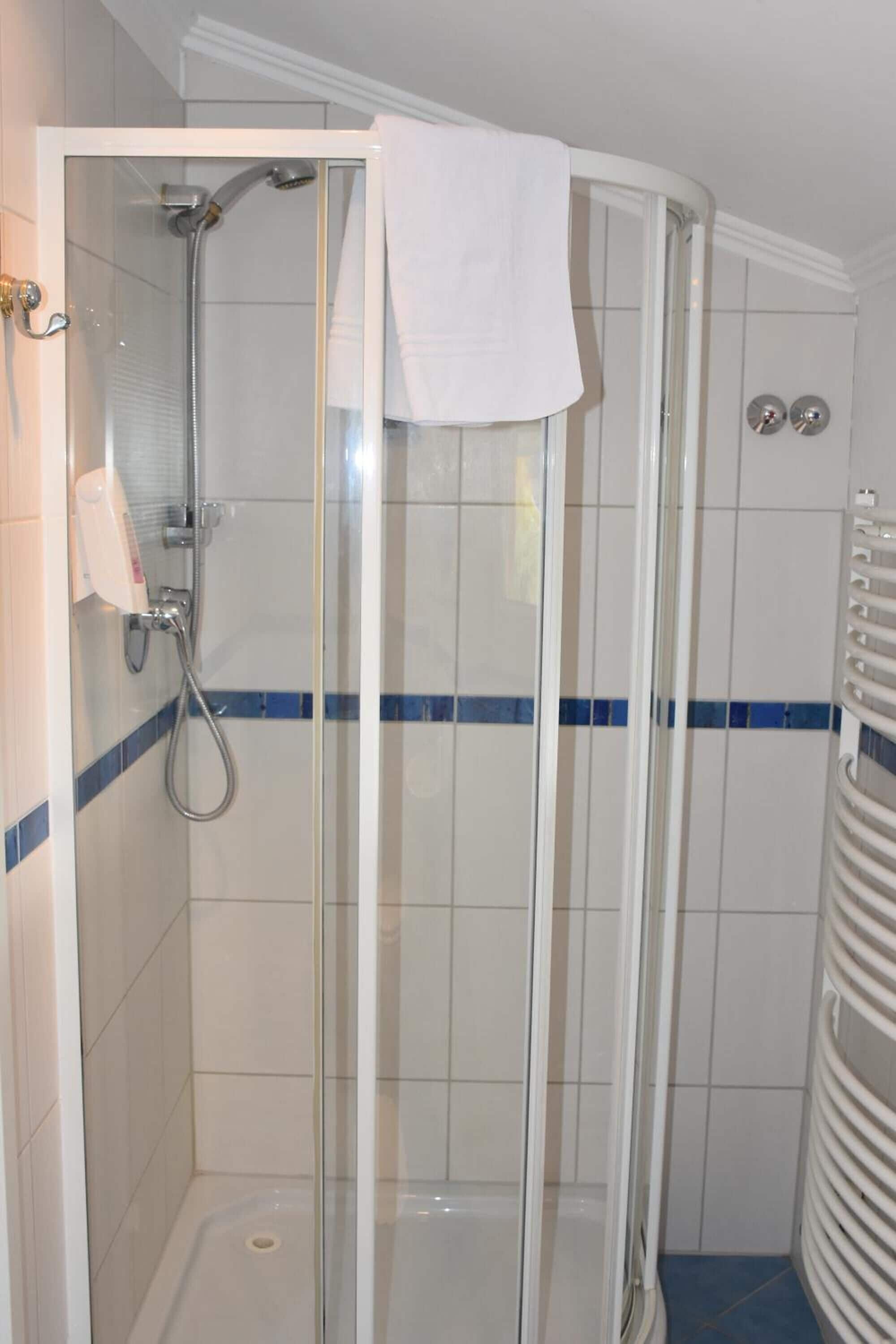 Un box de ducha con paredes de vidrio, un cabezal de ducha manual, una toalla blanca colgada en la puerta y un toallero blanco a la derecha.