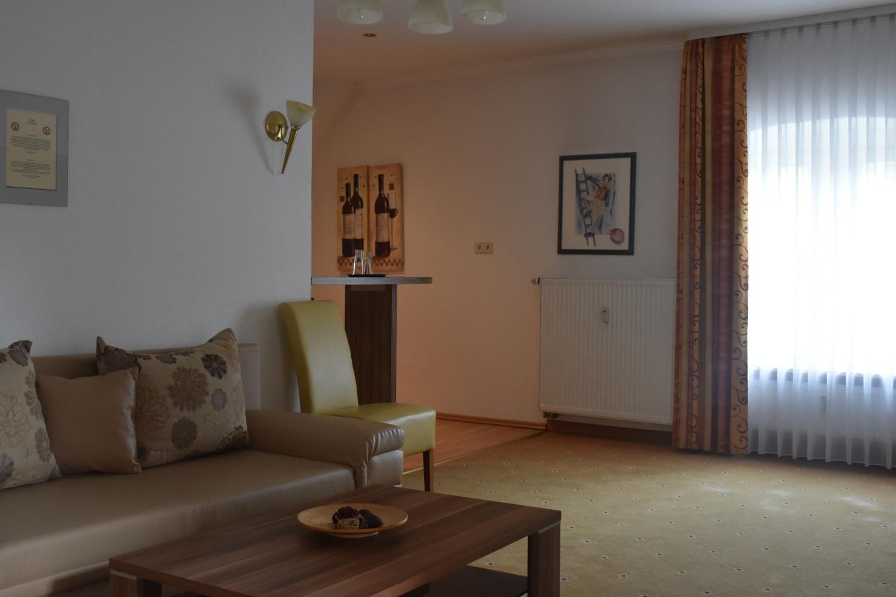 A living room with a sofa, a wooden coffee table, a chair, and a radiator. There is a framed picture on the wall and a window with curtains.