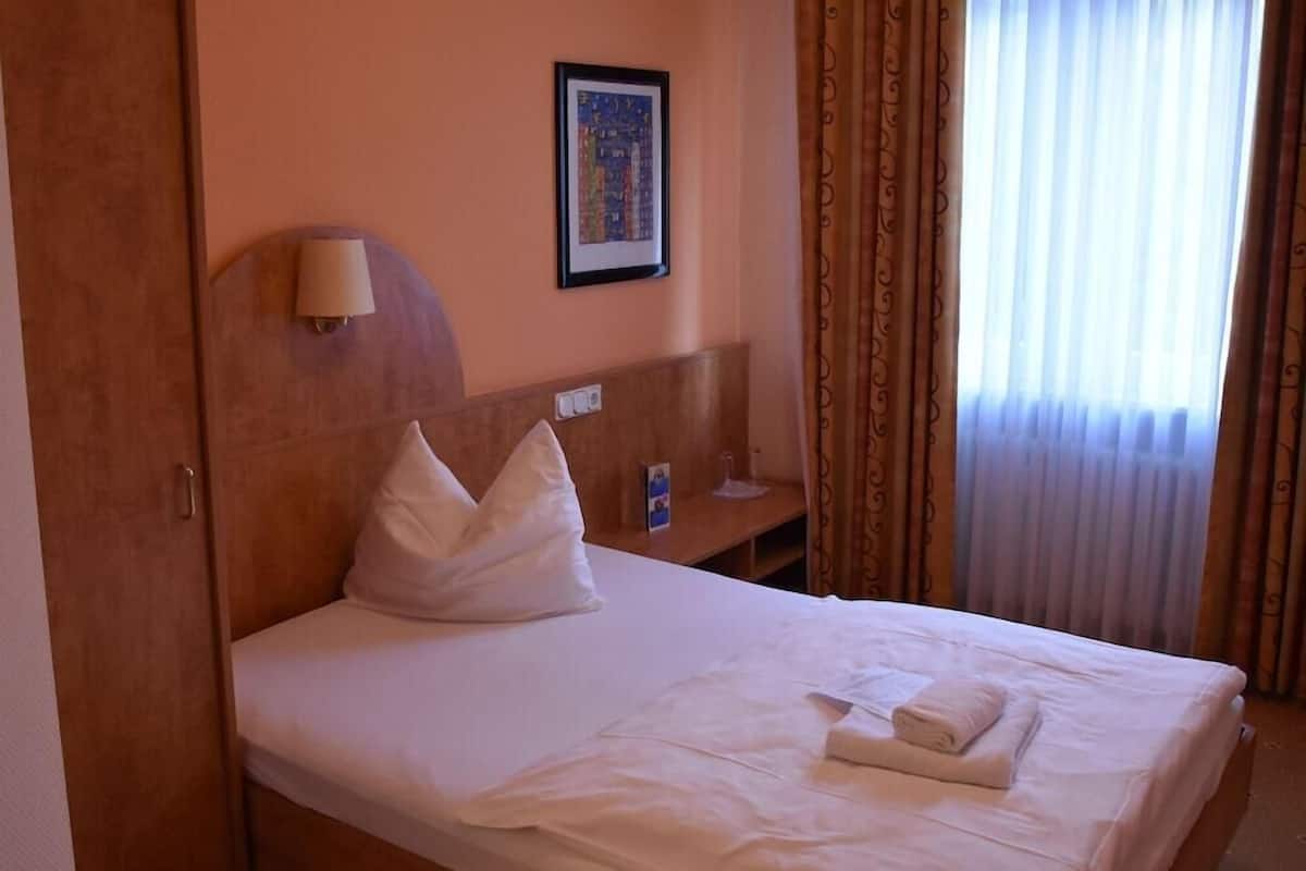 A hotel room with a bed, a wall-mounted lamp, a framed picture, a bedside table, and a window with curtains.