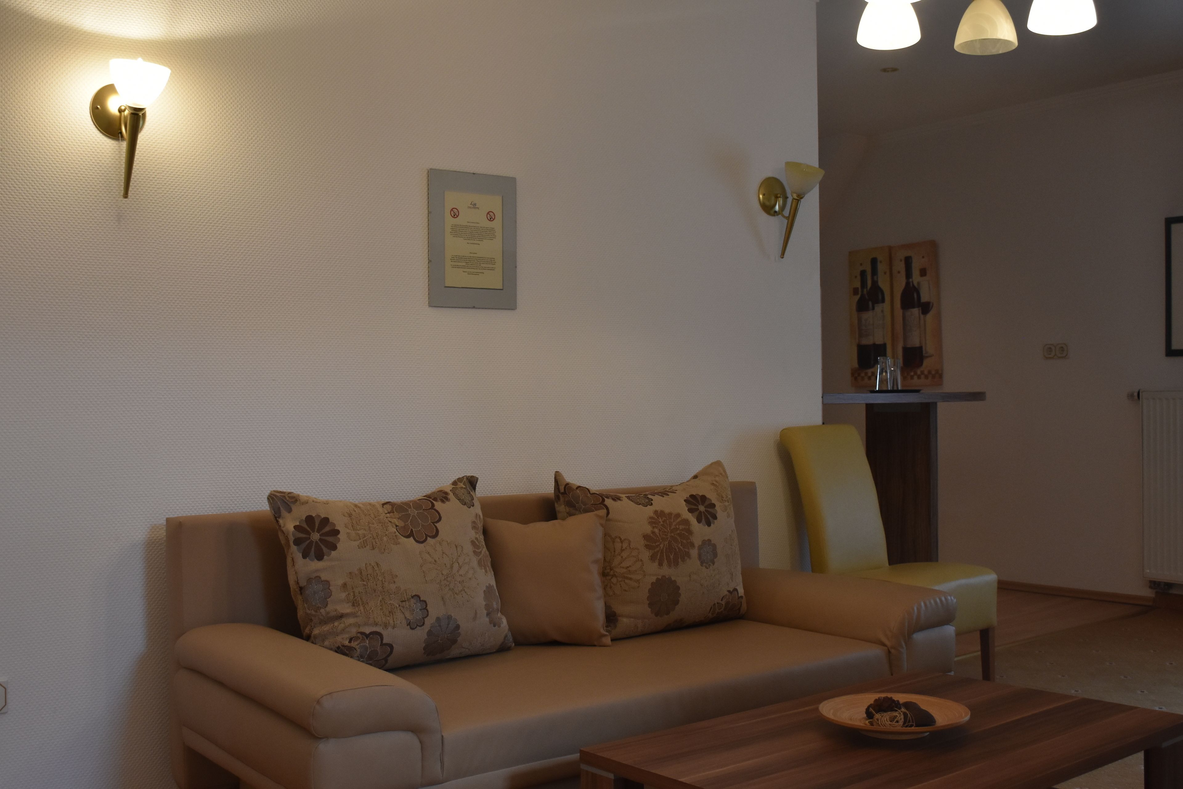 A living room with a sofa, a wooden coffee table, and a chair. There are wall-mounted lights and a framed certificate on the wall.